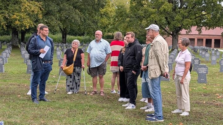 Mehrere Menschen mit traurigen Mienen stehen auf einem Rasenfeld voller Grabsteine, einer spricht. Mehrere Menschen mit traurigen Mienen stehen auf einem Rasenfeld voller Grabsteine, einer spricht.