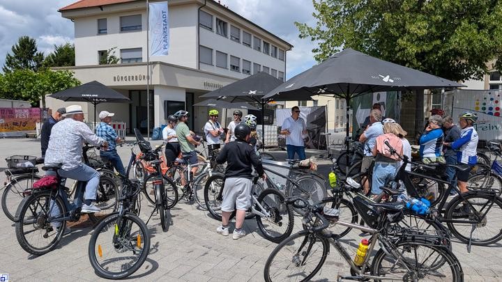 Unter und vor einem Sonnenschirm stehen viele Radfahrer und hören einem Mann zu, der mit ihnen spricht