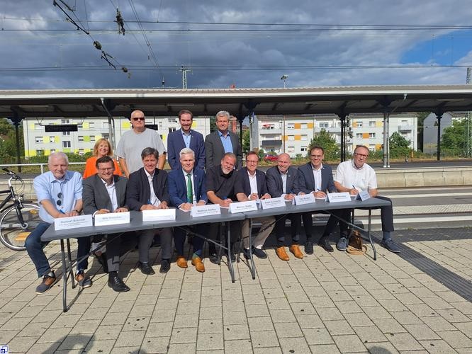 Viele Menschen sitzen an einem langen Tisch und unterzeichnen ein Dokument, vier Personen stehen dahinter